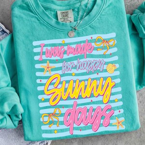 Pu&ograve; includere: Felpa Comfort Colors color turchese con la scritta "I was made for happy sunny days" in rosa e giallo. Il design include strisce blu, stelle marine, fiocchi e una conchiglia. &Egrave; visibile una collana d'argento.
