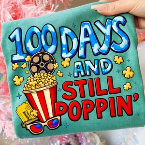Puede incluir: Camiseta de color turquesa con un gr&aacute;fico colorido. El dise&ntilde;o incluye el texto "100 DAYS AND STILL POPPIN'" en azul y rojo, junto con ilustraciones de palomitas de ma&iacute;z, una bobina de pel&iacute;cula, gafas 3D y una entrada de cine. Prenda casual.