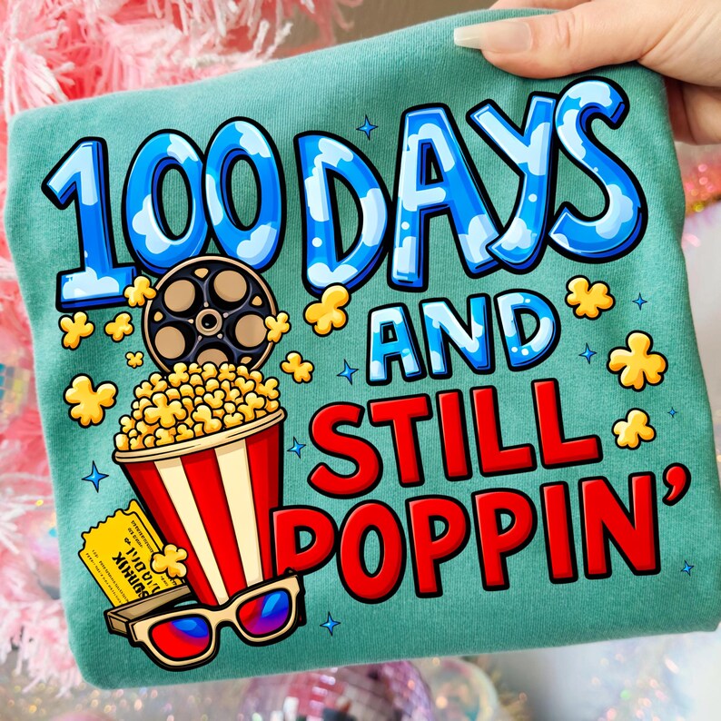 Puede incluir: Camiseta de color turquesa con un gr&aacute;fico colorido. El dise&ntilde;o incluye el texto "100 DAYS AND STILL POPPIN'" en azul y rojo, junto con ilustraciones de palomitas de ma&iacute;z, una bobina de pel&iacute;cula, gafas 3D y una entrada de cine. Prenda casual.