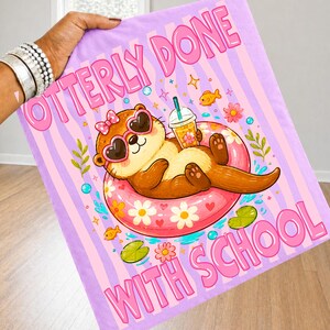 K&ouml;nnte beinhalten: Ein helllila und rosa Strandtuch mit dem Text "OTTERLY DONE WITH SCHOOL". Das Tuch zeigt einen Cartoon-Otter mit herzf&ouml;rmiger Sonnenbrille, der in einem rosa Schwimmring schwimmt und ein Getr&auml;nk h&auml;lt. Der Hintergrund hat rosa Streifen und florale Akzente.
