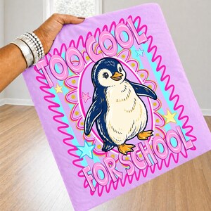 K&ouml;nnte beinhalten: Eine hellviolette Decke mit einem Cartoon-Pinguin und dem Text "TOO COOL FOR SCHOOL" in Pink. Der Pinguin ist blau, wei&szlig; und gelb, umgeben von einem dekorativen Design mit Sternen und Wellenlinien.
