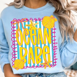 May include: Light blue sweatshirt with the words "OCEAN BABE" in yellow, framed by a pink border. The design includes yellow starfish and seashells on a striped background. The sweatshirt has long sleeves.