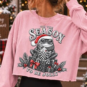 Puede incluir: Una sudadera corta de color rosa claro con el gr&aacute;fico de una rana con gorro de Pap&aacute; Noel y gafas, sosteniendo una taza de cacao sobre cajas de regalo. El texto dice TIS THE SEASON TO BE JOLLY, rodeado de hojas de acebo y bayas rojas.