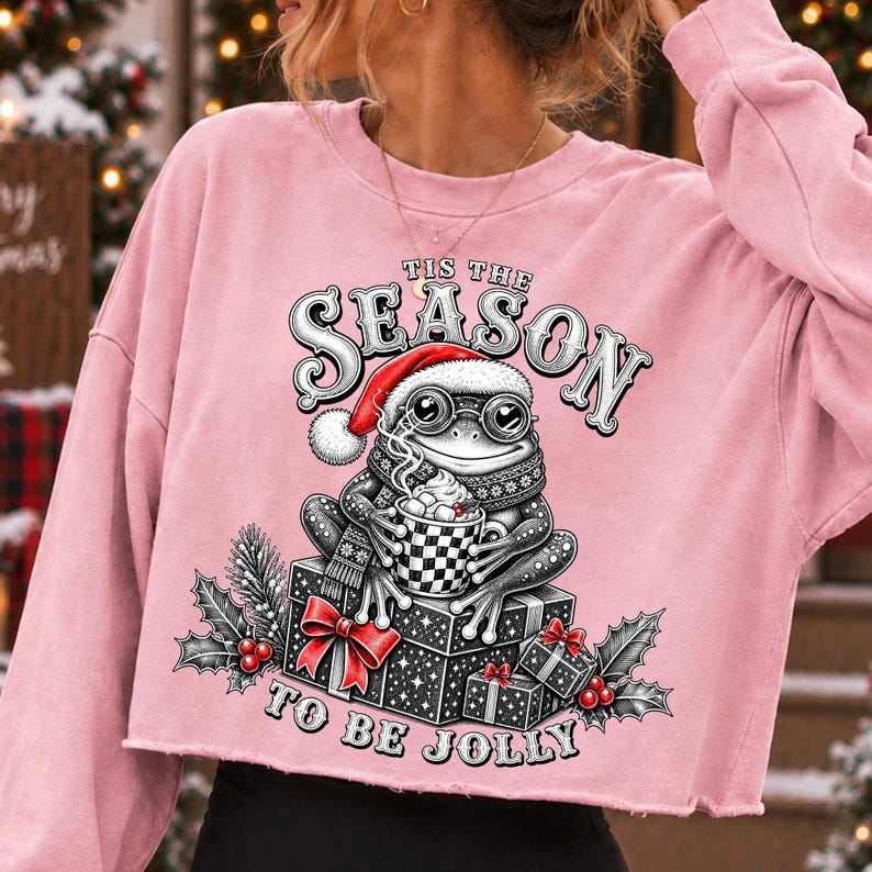 Puede incluir: Una sudadera corta de color rosa claro con el gr&aacute;fico de una rana con gorro de Pap&aacute; Noel y gafas, sosteniendo una taza de cacao sobre cajas de regalo. El texto dice TIS THE SEASON TO BE JOLLY, rodeado de hojas de acebo y bayas rojas.