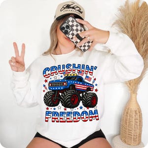 K&ouml;nnte beinhalten: Wei&szlig;es Sweatshirt mit einem blauen Monster-Truck-Motiv mit rot-wei&szlig;en Streifen, den Worten "CRUSHIN' FREEDOM" in Rot, Wei&szlig; und Blau und Sternen. Eine beige Baseballkappe und ein Telefon mit einer schwarz-wei&szlig; karierten H&uuml;lle sind ebenfalls zu sehen.