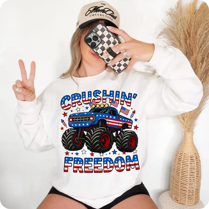 K&ouml;nnte beinhalten: Wei&szlig;es Sweatshirt mit einem blauen Monster-Truck-Motiv mit rot-wei&szlig;en Streifen, den Worten "CRUSHIN' FREEDOM" in Rot, Wei&szlig; und Blau und Sternen. Eine beige Baseballkappe und ein Telefon mit einer schwarz-wei&szlig; karierten H&uuml;lle sind ebenfalls zu sehen.