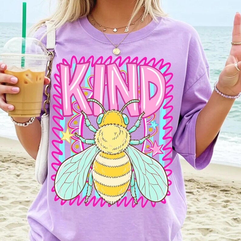 May include: A lilac-colored t-shirt with a large graphic of a bee and the word "KIND" in pink and blue. The bee is yellow and white with blue wings. The shirt has short sleeves.
