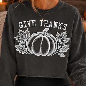 Puede incluir: Una sudadera corta de manga larga en color gris carb&oacute;n con un estampado gr&aacute;fico blanco de una calabaza rodeada de hojas de arce y roble. El texto "GIVE THANKS" est&aacute; impreso en una fuente blanca de estilo desgastado sobre la ilustraci&oacute;n.