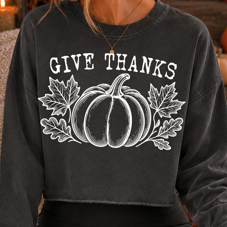 Puede incluir: Una sudadera corta de manga larga en color gris carb&oacute;n con un estampado gr&aacute;fico blanco de una calabaza rodeada de hojas de arce y roble. El texto "GIVE THANKS" est&aacute; impreso en una fuente blanca de estilo desgastado sobre la ilustraci&oacute;n.