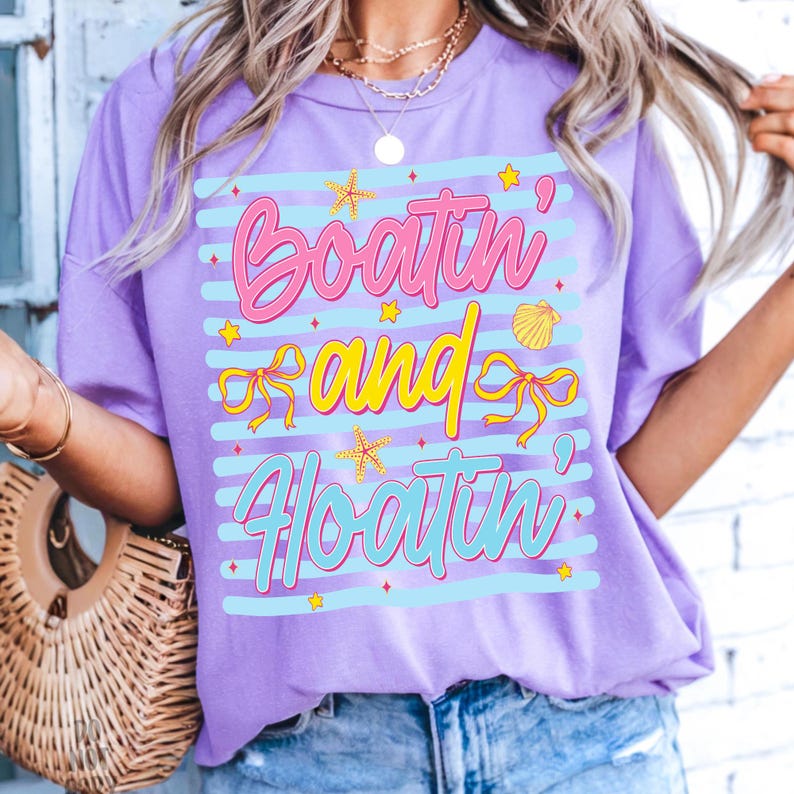 以下が含まれることがあります： ピンクと黄色の文字で「Boatin' and Floatin'」と書かれたライラック色のTシャツ。デザインには、ヒトデ、貝殻、リボンが青と白のストライプで描かれています。カジュアルでリラックスしたフィット感のシャツです。