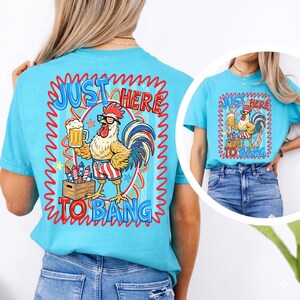 May include: A light blue t-shirt featuring a cartoon rooster wearing sunglasses and red, white, and blue striped shorts, holding a beer. The shirt has the text "JUST HERE TO BANG" in a red and blue wavy design.