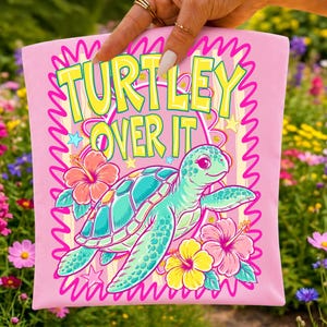 Puede incluir: Camiseta rosa con un dise&ntilde;o de tortuga de dibujos animados. La tortuga es verde azulado y turquesa, rodeada de flores de hibisco rosas, amarillas y rojas. El texto "TURTLEY OVER IT" est&aacute; en amarillo y verde.