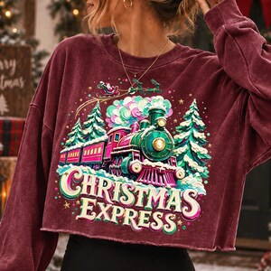 May include: A maroon long-sleeve crewneck sweatshirt featuring a graphic print of a green and red steam locomotive train, snow-covered pine trees, and a flying sleigh. The text "CHRISTMAS EXPRESS" is printed in large, bold, cream-coloured letters.