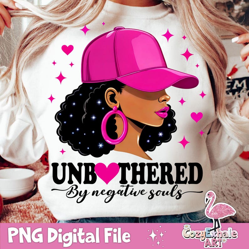 May include: A white sweatshirt with a pink and black graphic design. The graphic features a woman with black hair wearing a pink baseball cap and large pink earrings. The text "Unbothered by negative souls" is written in black with a pink heart in the middle of the word "unbothered".