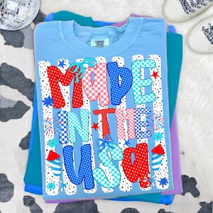 Pode incluir: Uma t-shirt azul-clara com o texto MADE IN THE USA numa fonte estampada multicolorida. As letras est&atilde;o decoradas com bolinhas e padr&otilde;es de xadrez vermelhos, brancos e azuis. O gr&aacute;fico inclui fogo de artif&iacute;cio e detalhes de la&ccedil;os azuis.