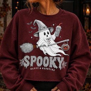 May include: A maroon long-sleeve sweatshirt featuring a graphic of a ghost wearing a witch hat, riding a broomstick, and holding a drink. The ghost is accompanied by bats and a jack-o'-lantern. The text reads SPOOKY and HAPPY HAUNTING.
