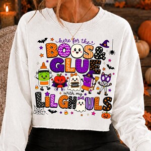 K&ouml;nnte beinhalten: Ein wei&szlig;es Langarm-Sweatshirt mit einer bunten Halloween-Grafik und dem Text "here for the BOOS & GLUE with my LIL GHOULS". Das Design zeigt Zeichentrickfiguren wie einen Geist, eine Klebeflasche, einen Apfel und eine Katze sowie Flederm&auml;use und Sterne.