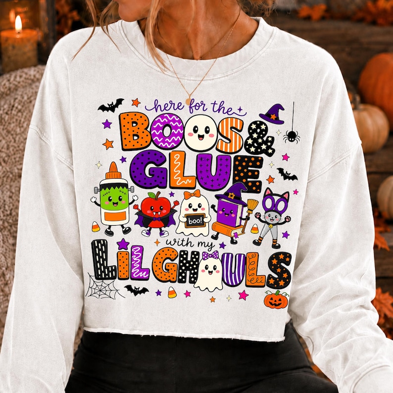K&ouml;nnte beinhalten: Ein wei&szlig;es Langarm-Sweatshirt mit einer bunten Halloween-Grafik und dem Text "here for the BOOS & GLUE with my LIL GHOULS". Das Design zeigt Zeichentrickfiguren wie einen Geist, eine Klebeflasche, einen Apfel und eine Katze sowie Flederm&auml;use und Sterne.