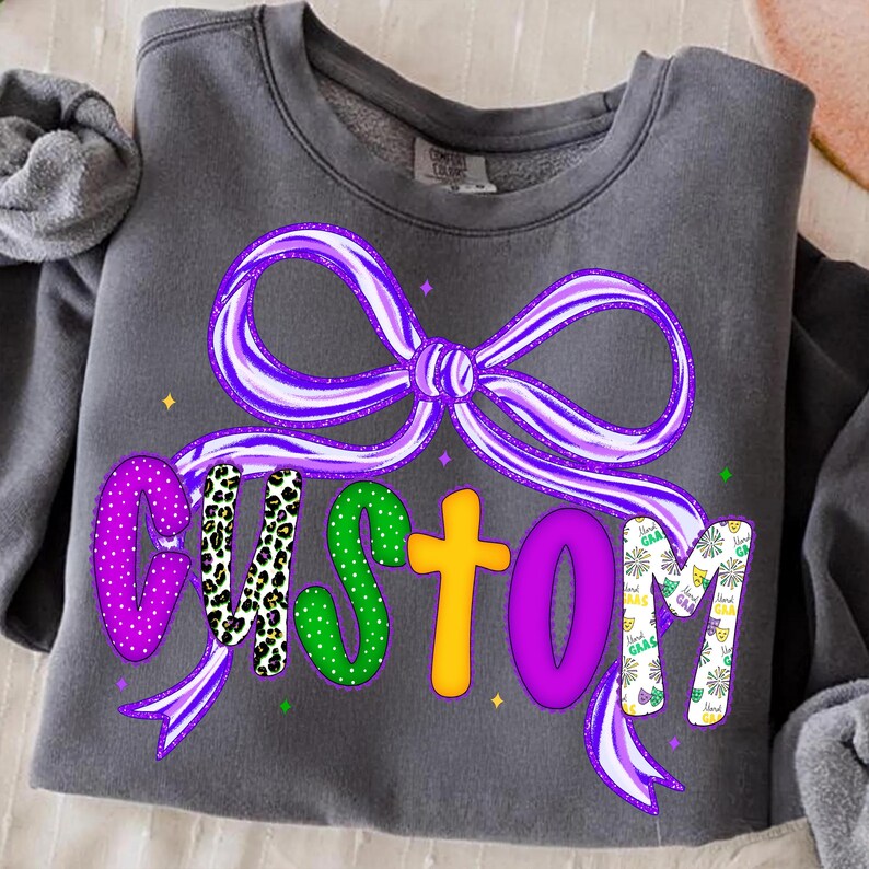 May include: Dark gray sweatshirt with a large purple bow and the word "CUSTOM" in colorful, patterned letters. The letters are decorated with polka dots, leopard print, and Mardi Gras-themed designs.
