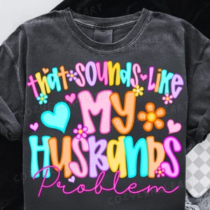 K&ouml;nnte beinhalten: Dunkelgraues T-Shirt mit dem Schriftzug "that sounds like my husbands problem" in bunten Buchstaben. Das Design beinhaltet Herzen und Blumen in Blau-, Rosa-, Gelb- und Oranget&ouml;nen.