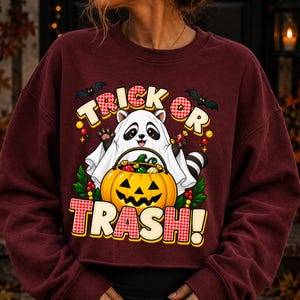 May include: A maroon long-sleeve sweatshirt featuring a graphic of a raccoon wearing a white ghost sheet, holding a jack-o'-lantern bucket filled with sweets. The text reads TRICK OR TRASH in a red and white chequered font with black bats.