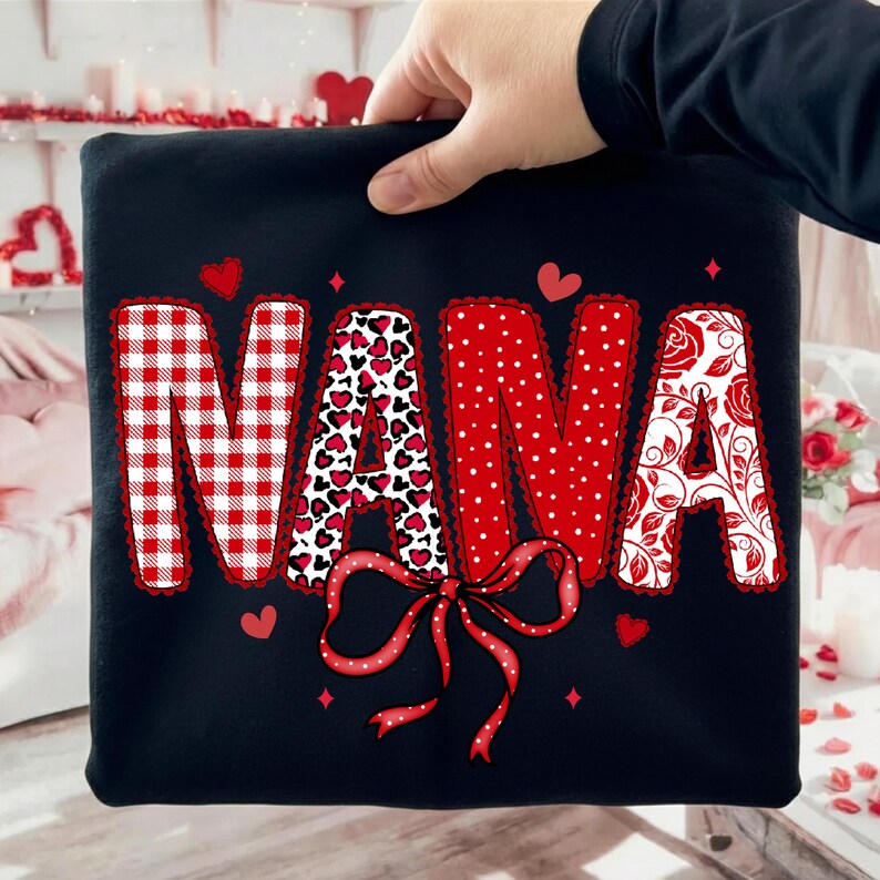 Może przedstawiać: Czarna bluza z napisem "NANA" w ozdobnej czcionce. Litery mają r&oacute;żne wzory, w tym czerwono-białą kratę, serca w panterkę, kropki i kwiatowe wzory. Na dole znajduje się czerwona kokarda.