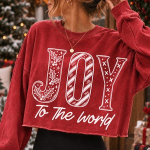 May include: A red long-sleeve cropped sweatshirt featuring the white text JOY with holly leaves, candy cane stripes, and cross patterns inside the letters. Below the word JOY, the white text reads To The world in a cursive script font.