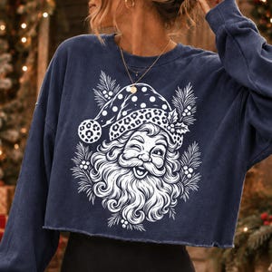 K&ouml;nnte beinhalten: Ein marineblaues, lang&auml;rmeliges bauchfreies Sweatshirt mit einem wei&szlig;en Grafikdruck eines zwinkernden Weihnachtsmanns, der eine M&uuml;tze mit Leopardenmuster tr&auml;gt. Das Design ist von Tannenzweigen und Stechpalmenbeeren umgeben.