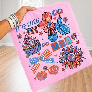 Pode incluir: Um cobertor rosa com desenhos patri&oacute;ticos em vermelho, branco e azul. O cobertor tem um cupcake, animais de bal&atilde;o, cerejas e uma flor. O texto "USA" e "1776-2026" tamb&eacute;m s&atilde;o exibidos.