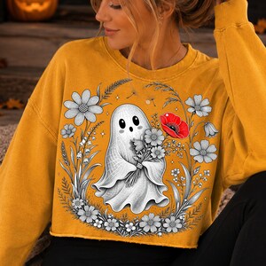 K&ouml;nnte beinhalten: Ein senfgelbes Langarm-Sweatshirt mit einem Grafikdruck eines wei&szlig;en Geistes, der einen Blumenstrau&szlig; h&auml;lt. Das Design zeigt schwarz-wei&szlig;e Strichzeichnungen von Blumen um den Geist herum, erg&auml;nzt durch eine einzelne leuchtend rote Mohnblume.