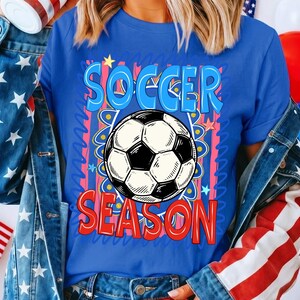 Puede incluir: Una camiseta azul con las palabras "SOCCER SEASON" en un dise&ntilde;o colorido, con un gr&aacute;fico de bal&oacute;n de f&uacute;tbol. La camiseta tiene cuello redondo y mangas cortas. El dise&ntilde;o incluye detalles en rojo, azul y amarillo.
