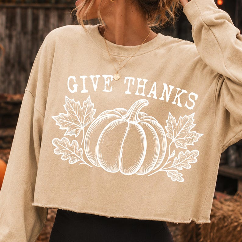 Puede incluir: Una sudadera corta de color beige con mangas largas que presenta un estampado gr&aacute;fico blanco de una calabaza rodeada de hojas de arce y roble. El texto "GIVE THANKS" est&aacute; impreso en una fuente serif blanca sobre la ilustraci&oacute;n.