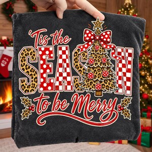 Op de afbeelding: Een donkergrijs t-shirt met de tekst 'Tis the SEASON to be Merry' in rood en wit. Het ontwerp bevat een kerstboom met luipaardprint, rood-witte stippen als versiering en een grote rood-wit gestippelde strik aan de bovenkant.
