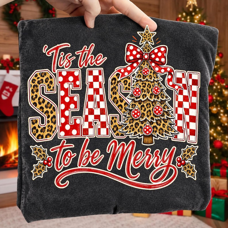 Op de afbeelding: Een donkergrijs t-shirt met de tekst 'Tis the SEASON to be Merry' in rood en wit. Het ontwerp bevat een kerstboom met luipaardprint, rood-witte stippen als versiering en een grote rood-wit gestippelde strik aan de bovenkant.
