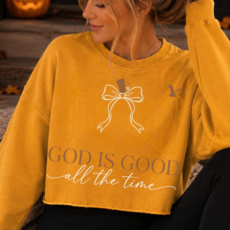 Puede incluir: Una sudadera corta de manga larga en color amarillo mostaza con un gr&aacute;fico de l&iacute;neas blancas de una calabaza con un lazo encima. El texto "GOD IS GOOD" est&aacute; impreso en una fuente oscura y negrita, con "all the time" debajo en cursiva blanca.