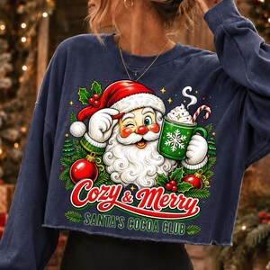 K&ouml;nnte beinhalten: Ein marineblaues Langarm-Sweatshirt mit einer Grafik von einem zwinkernden Weihnachtsmann, der einen gr&uuml;nen Kakaobecher mit Zuckerstange h&auml;lt. Der Text lautet Cozy & Merry Santa's Cocoa Club, umgeben von Weihnachtsb&auml;umen, Stechpalmen und Kugeln.