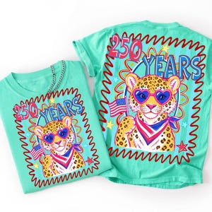 Op de afbeelding: Twee turquoise t-shirts met een afbeelding van een luipaard met een hartvormige zonnebril en een Amerikaanse vlag. Het ontwerp bevat de tekst "250 YEARS" in rood en blauw, omgeven door een golvende rode rand en sterren.