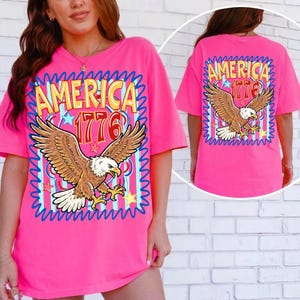 May include: A bright pink t-shirt featuring a graphic of an eagle with outstretched wings. The text "AMERICA 1776" is displayed above the eagle. The design is framed by a blue wavy border. The back of the shirt also displays the same design.