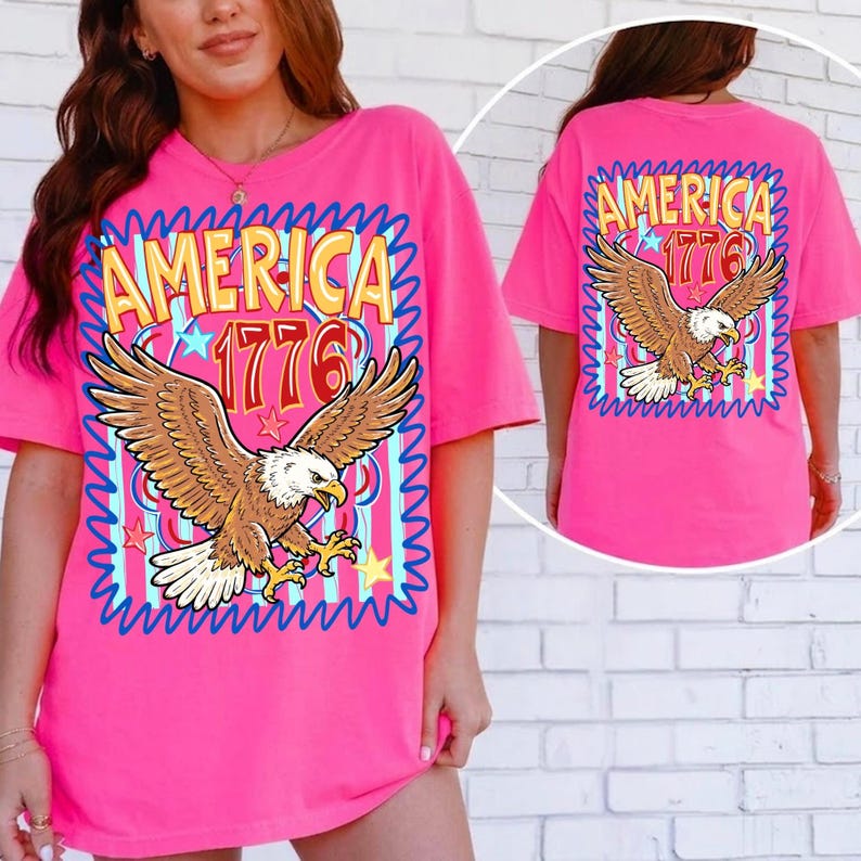 May include: A bright pink t-shirt featuring a graphic of an eagle with outstretched wings. The text "AMERICA 1776" is displayed above the eagle. The design is framed by a blue wavy border. The back of the shirt also displays the same design.