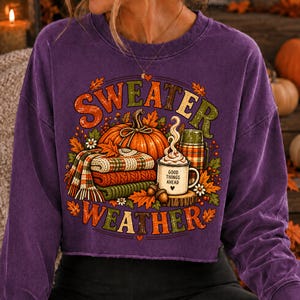 Puede incluir: Una sudadera morada de manga larga con un estampado gr&aacute;fico que incluye el texto "SWEATER WEATHER". El dise&ntilde;o muestra una calabaza naranja grande, una pila de mantas a cuadros dobladas, un termo y una taza con el texto "GOOD THINGS AHEAD".