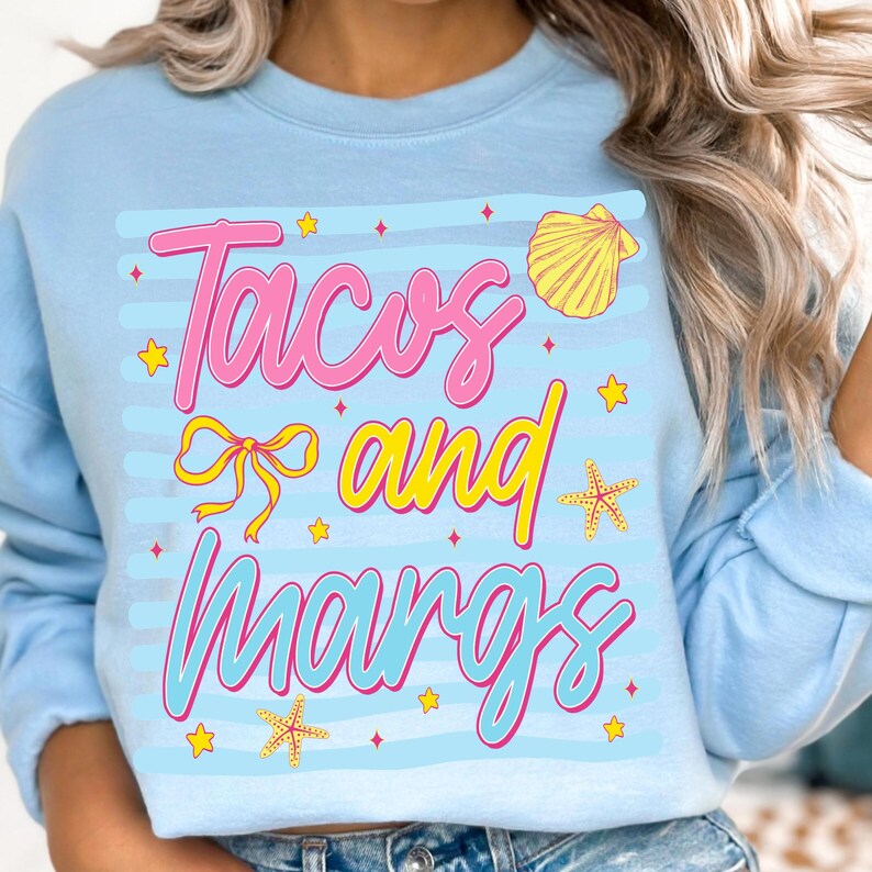 May include: Pale blue sweatshirt featuring the text "Tacos and Margs" in pink and yellow. The design includes a yellow seashell, starfish, and stars. A casual, comfortable style of sweatshirt.