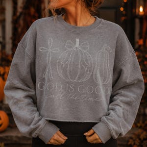 K&ouml;nnte beinhalten: Ein graues Sweatshirt mit Rundhalsausschnitt und einem wei&szlig;en Glitzer-Aufdruck, der ein Kreuz, einen K&uuml;rbis mit Schleife und einen Flaschenk&uuml;rbis zeigt. Der Text "GOD IS GOOD" ist fett gedruckt, darunter steht "all the time" in Schreibschrift.