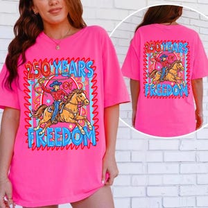 Puede incluir: Camiseta rosa brillante con un gr&aacute;fico de un vaquero montando a caballo, con el texto "250 YEARS OF FREEDOM". El dise&ntilde;o est&aacute; enmarcado por un borde ondulado azul y rojo. La parte trasera de la camiseta tambi&eacute;n tiene el mismo gr&aacute;fico.