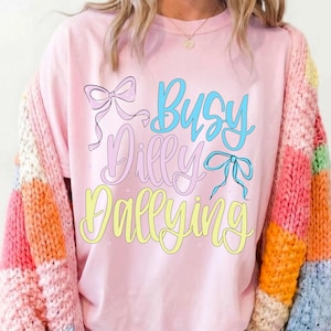 Puede incluir: Una camiseta rosa claro con la frase "Busy Dilly Dallying" en escritura azul, rosa y amarilla, adornada con lazos. La camiseta se combina con un c&aacute;rdigan a rayas de colores.
