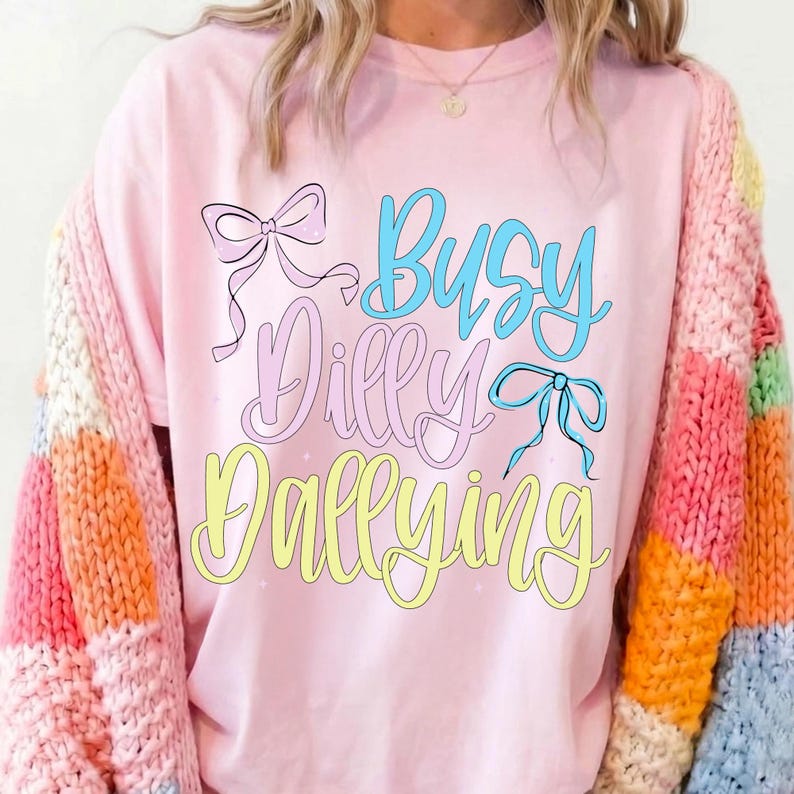 Puede incluir: Una camiseta rosa claro con la frase "Busy Dilly Dallying" en escritura azul, rosa y amarilla, adornada con lazos. La camiseta se combina con un c&aacute;rdigan a rayas de colores.