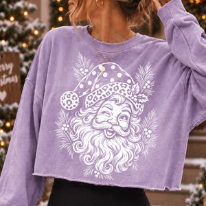 K&ouml;nnte beinhalten: Ein hellviolettes, lang&auml;rmeliges bauchfreies Sweatshirt mit einem wei&szlig;en Grafikdruck eines zwinkernden Weihnachtsmanns, der eine M&uuml;tze mit Leopardenmuster und Punkten tr&auml;gt, umgeben von Tannennadeln und Stechpalmenbeeren auf der Vorderseite.