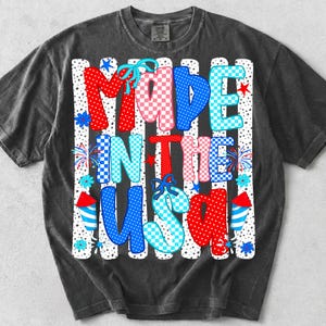 Pode incluir: Uma t-shirt cinzento-carv&atilde;o de manga curta com uma estampa gr&aacute;fica com o texto MADE IN THE USA. As letras s&atilde;o preenchidas com padr&otilde;es xadrez e de bolinhas em vermelho, branco e azul, acentuadas por pequenos la&ccedil;os azuis e estrelas.
