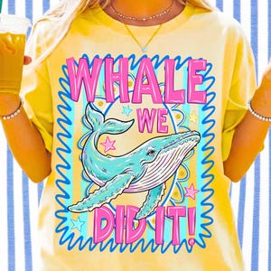 K&ouml;nnte beinhalten: Gelbes T-Shirt mit einer Illustration eines blauen Wals und dem Text "WHALE WE DID IT!" in Pink und Blau. Das Design beinhaltet Sterne und einen dekorativen Rahmen, was einen verspielten Look erzeugt.