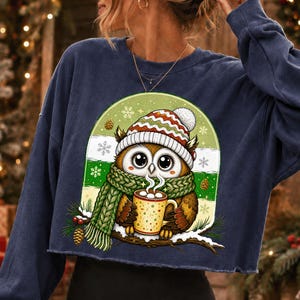 May include: A navy blue long-sleeve sweatshirt featuring a graphic of a brown owl wearing a red, white, and green striped knit hat and a green knit scarf. The owl holds a yellow mug with steam, set against a green arch with snowflakes.