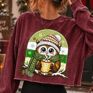 May include: A maroon long-sleeve crewneck sweatshirt featuring a graphic of an owl wearing a knit hat and scarf while holding a mug of hot cocoa. The owl is perched on a snowy branch against a green and white background with snowflake accents.